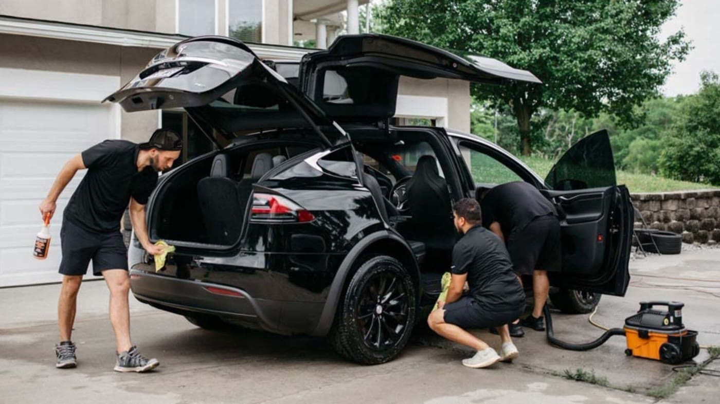Three people cleaning a black hatchback in a driveway, doors and hatch open
