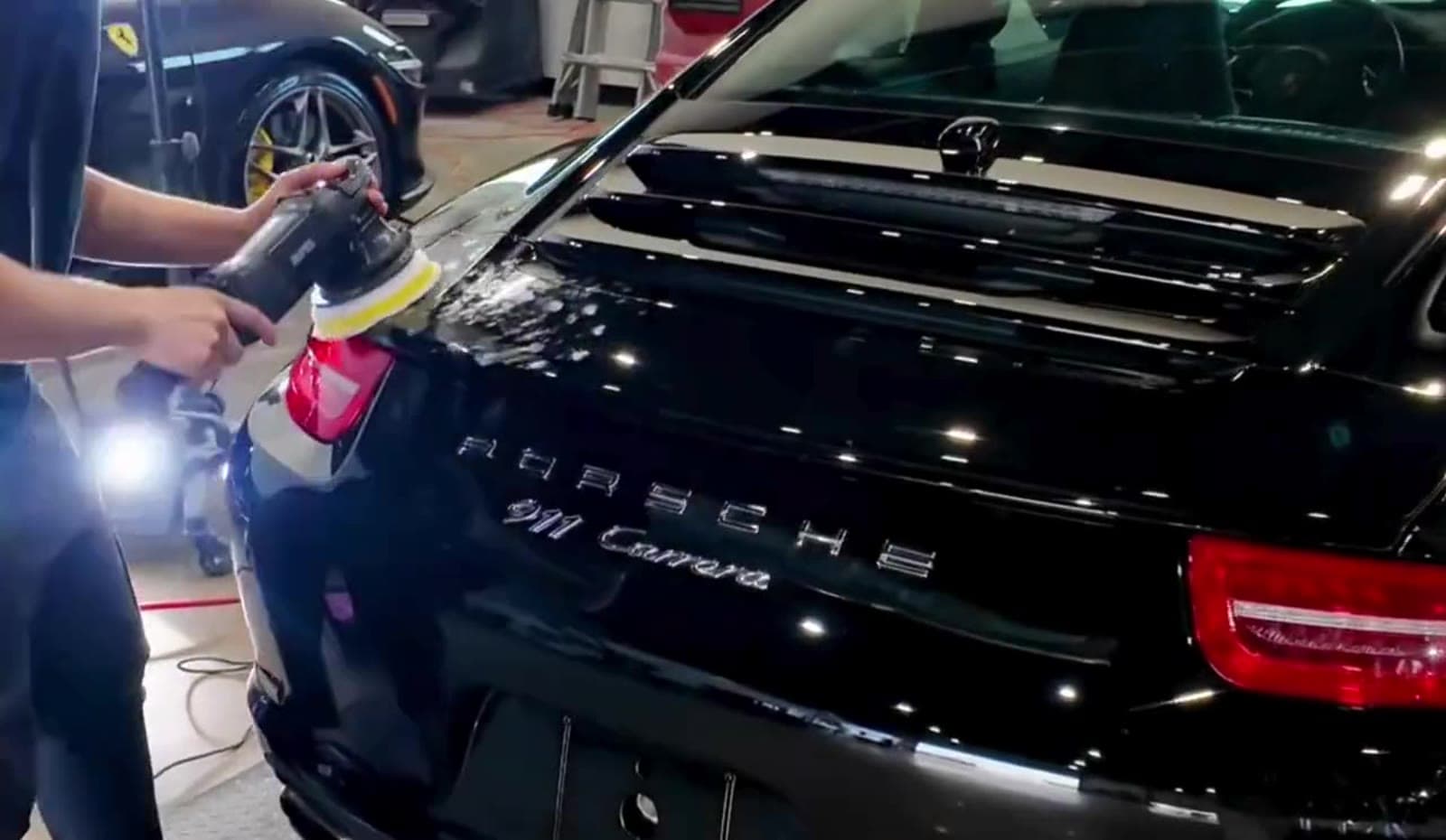 A detailer polishing the rear deck of a black Porsche 911 Carrera in a shop.